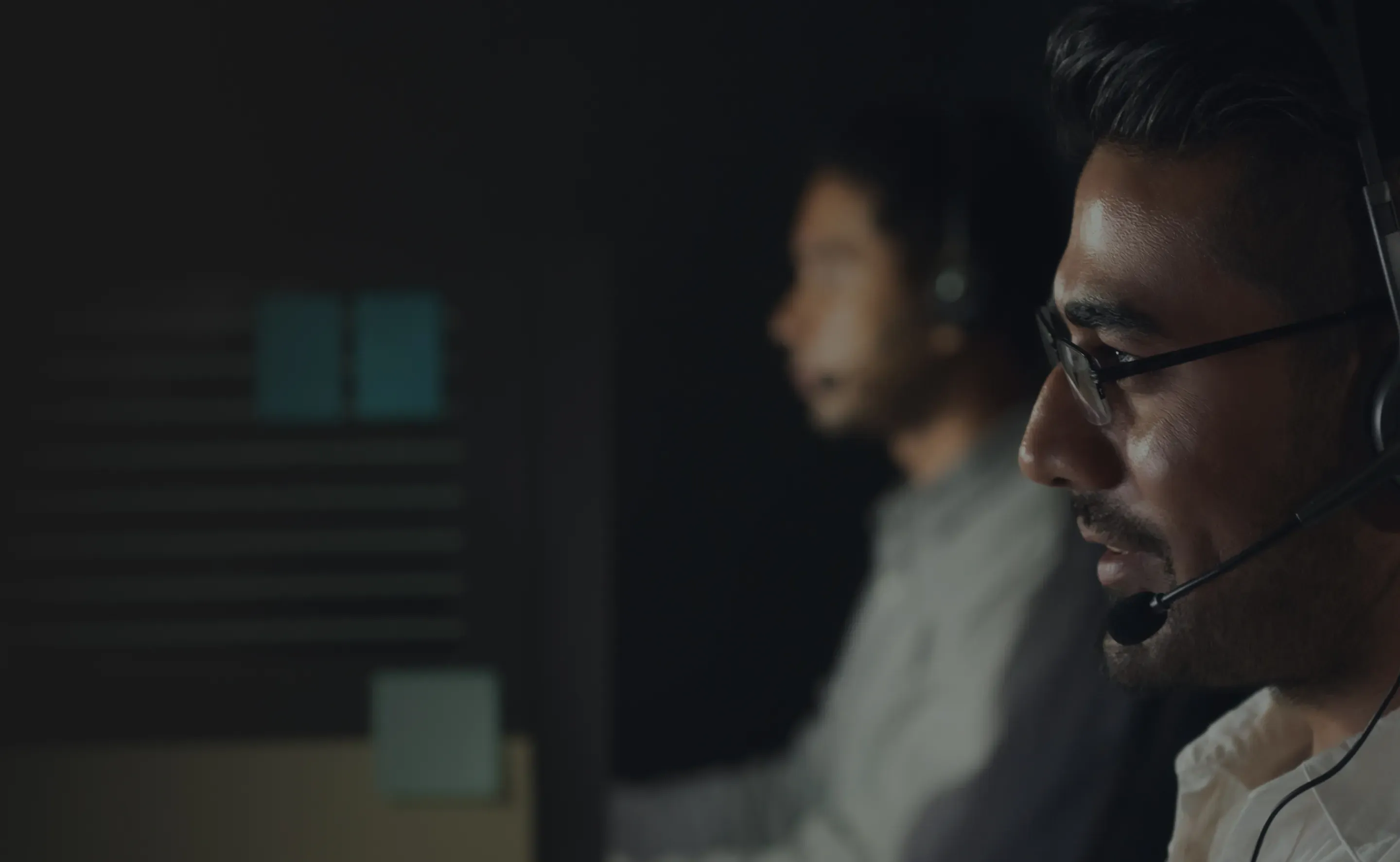 Contact center agent in glasses on a headset call, with a colleague working behind